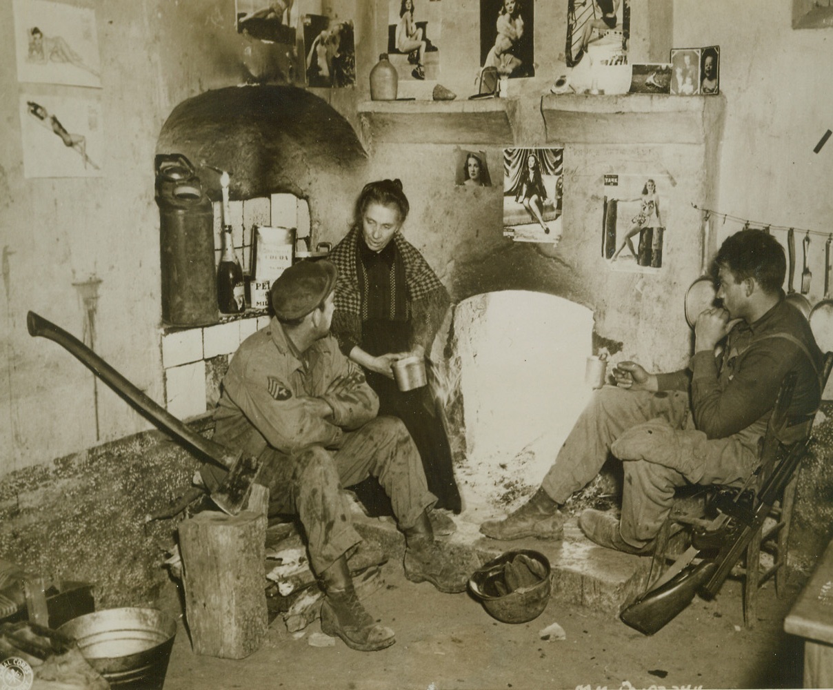 “Home, Sweet Home” in Italy, 4/3/1944. ITALY – Sgt. Vernon Dennis, (left), of Northome, Minn., and Pvt. Harry Boone, (right), of Oneonta, N.Y., warm themselves before the fire in their “home away from home” – the mayor’s house in a tiny Italian village, where the two members of a U.S. Army Photographic unit are quartered. The mayor’s wife, (center), has done her best to make the lads comfortable and has even allowed them to display their favorite collection of pinup girls on the walls and over the fireplace.Credit Line (U.S. Army Official Photo from Acme);
