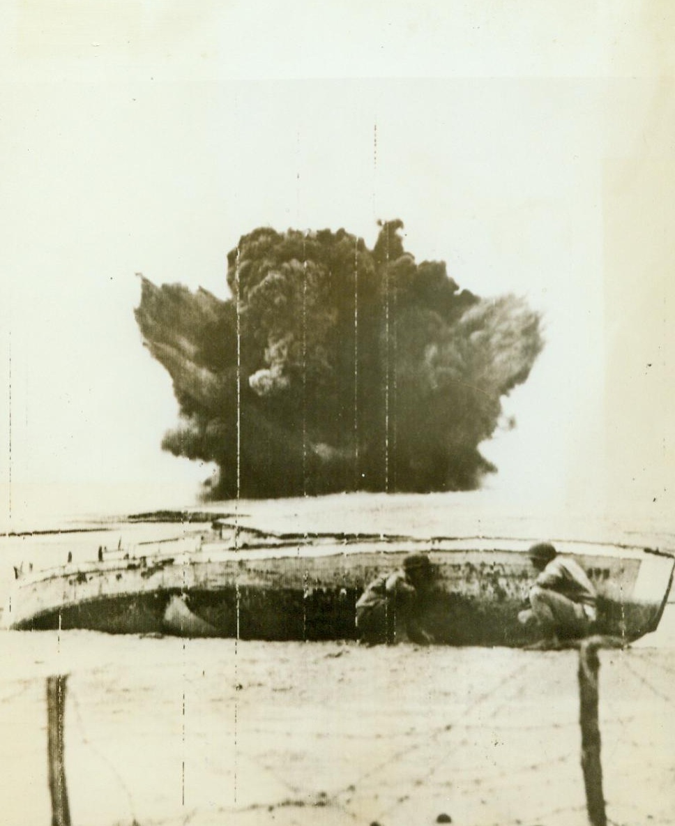 Any Shelter In Port Of Storm, 4/11/1944. Italy – Two American engineers use a derelict boat as protection from shrapnel as German teller mines explode in background. Sappers are making beachheads safe from German mines and barbed wire. Credit Line (US Army Radiotelephoto From ACME) 4/11/44;