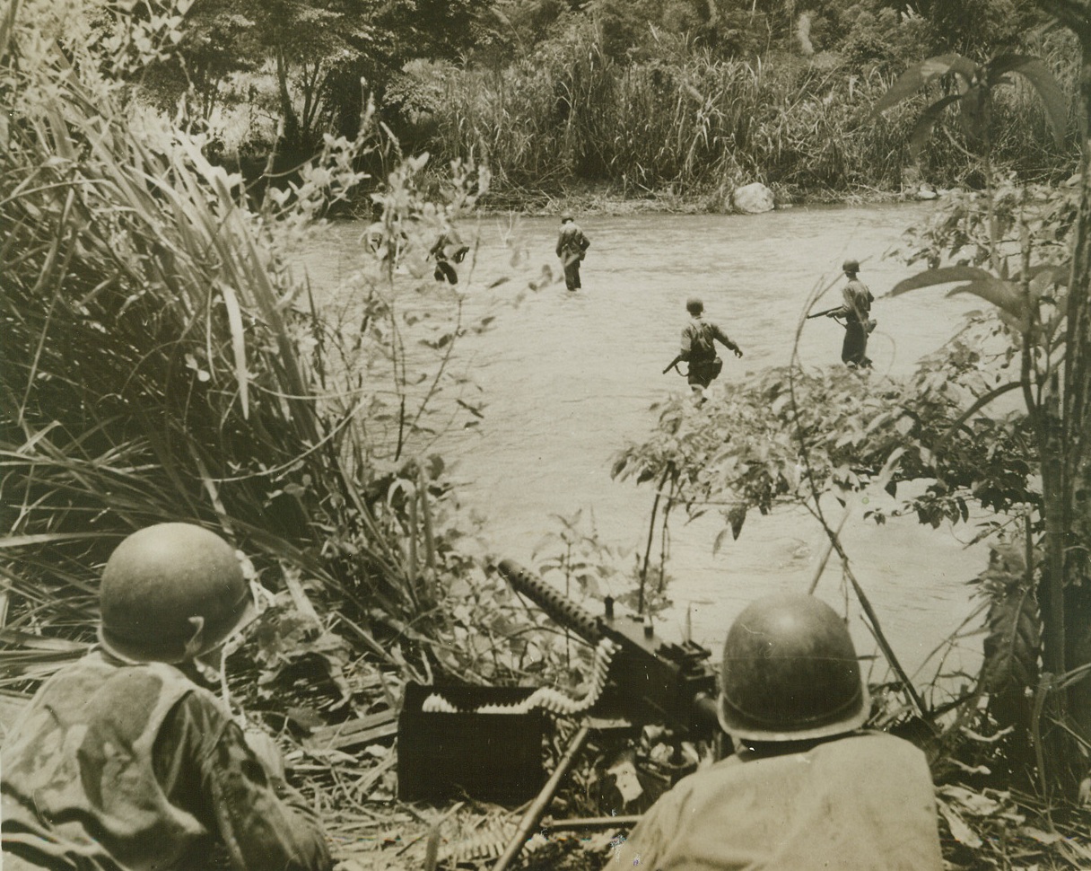 Advancing Under Cover, 4/9/1944. New Guinea – Cautiously advancing under the cover of a .50 caliber machine gun (foreground) American infantrymen wade across a river running through thick New Guinea jungle.  The heavy foliage rising immediately on the opposite side of the river could provide excellent hiding places for Jap snipers. Credit line (Signal Corps photo from ACME);