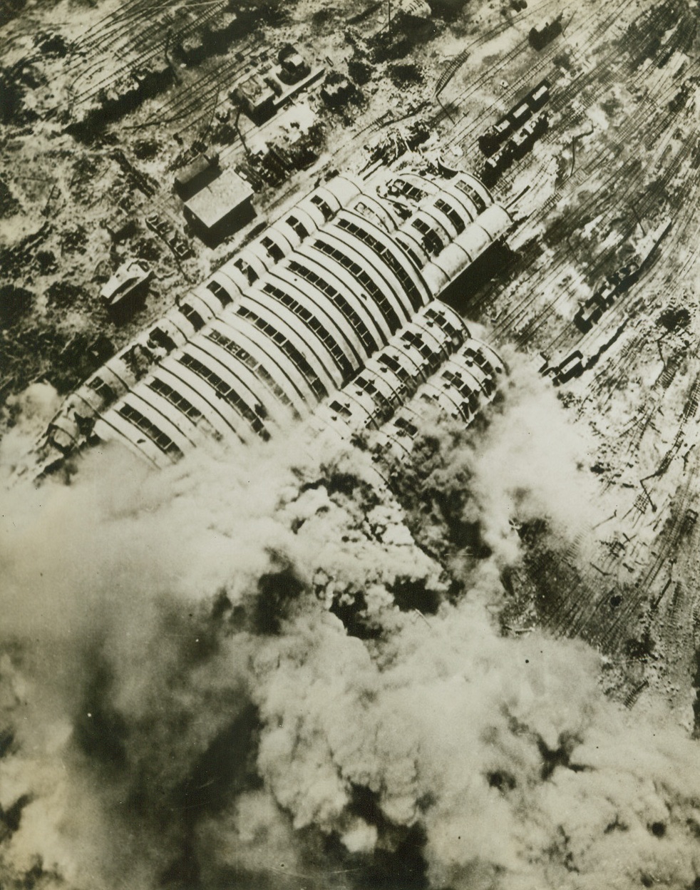 Damage Courtesy of the Allies, 4/29/1944. France – The R.A.F. bomber command recently dispatched more than 1,000 aircraft on a large number of operations, and in particular the offensive was resumed against railroad targets in France. Extensive damage was done to the railroad yards at Hirson in France. Clouds of smoke can be seen in this photo, taken at low level, covering the locomotive barns and repair shops. Many of the rail tracks leading into the barns and shops are seen to be wrecked, as well as freight cars near the shops. Credit: ACME;