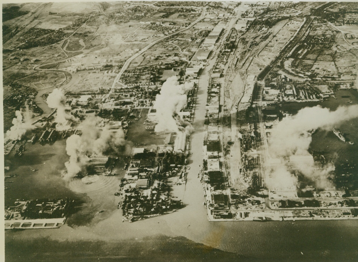 SOERABAYA DOCK AREA HIT, 5/30/1944. SOERABAYA, JAVA – Clouds of smoke billow up from docks and buildings as bombs crash down on the Jap-occupied town in Java. This scene was photographed during the recent large-scale Allied bombing raid on Soerabaya, when several important Japanese installations were hit, including machine shops, railroad yards, and power plant supplying the town with electricity. Credit: Official U.S. Navy photo from ACME;