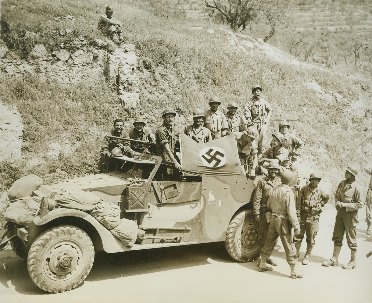 Sweet are Fruits of Revenge!, 5/23/1944. ESPERIA, ITALY—Smiling broadly, French infantry-men, who had seen their beloved country overrun by the Nazi horde, hold up a German flag they captured earlier in the day in the fighting reconnaissance car, and all of them wear U.S. equipment—all but their helmets, which they have retained as a badge of membership in the once-powerful Army of France.  Credit: ACME PHOTO BY CHARLES SEAWOOD FOR THE WAR PICTURE POOL.;