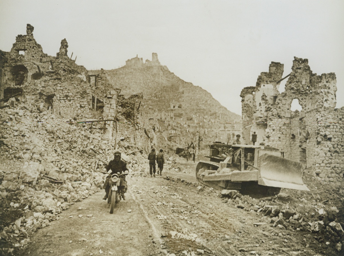 The Aftermath, 5/26/1944. CASSINO, ITALY—Allied bulldozers clear the road between the mountains of rubble that was once Cassino, as Army engineers work to repair the highway leading to Rome. The hotel des Roses, which housed German gun emplacements during the Allied siege, lies on the right, a mass of shattered wreckage. Hangman’s Hill, with the Benedictine Monastery atop it, lies in the background. All of the city, with the exception of the Monastery, has been declared “dead” by the Allies and no efforts are being made to rehabilitate it. Credit: ACME.;