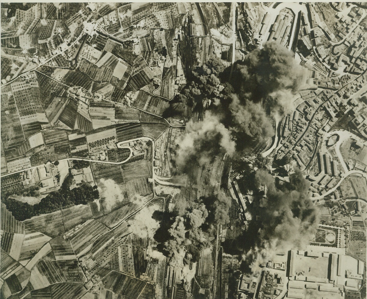 Ploesti Smashed Again by U.S. Airmen, 5/6/1944. PLOESTI, ROMANIA—Stepping up their non-stop pace in air offense, the Allies again hit the railroad marshalling yards at Ploesti and here Allied bombers wing over their target, leaving explosions and fires in their wake. The Flying Forts, based in Italy, were playing an early return engagement on the oil center which they attacked heavily not long ago. It appears that the Allies’ aim is to cripple German transportation and supply facilities as much as possible before the dawn of “D” day.  Credit: OFFICIAL AAF PHOTO FROM ACME.;