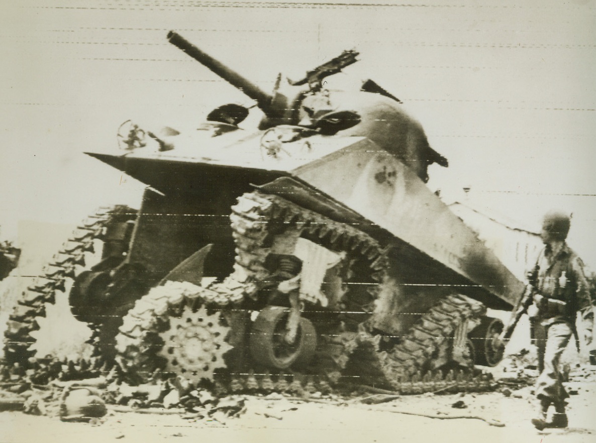 SET BACK ON ITS HEELS, 5/25/1944. ITALY—This American M-4 tank, disabled by a German Teller mine during the renewed offensive on the Anzio beachhead, near Borgo Sabotino, was blasted “back on its haunches” by a demolition charge set by an American patrol so that the monster wouldn’t prove of use to the enemy. Here, an American infantryman looks over the wrecked tank. Credit: Army Radiotelephoto from Acme;