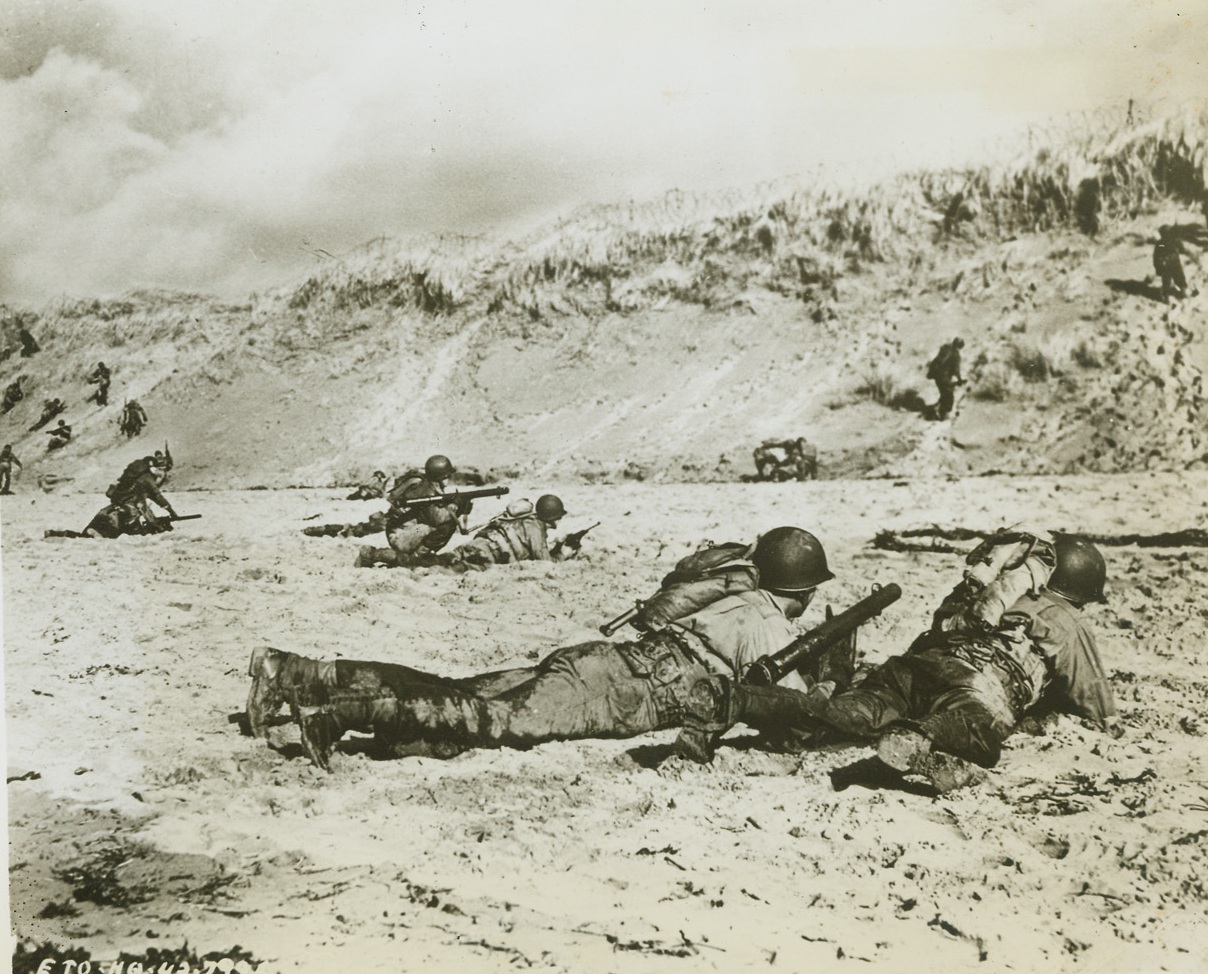 Up in Arms, 5/5/1944. England – “Hitting the deck” after landing on the beachhead, these American infantrymen train their MI Launcher Rockets, better known as “Bazookas,” on imaginary enemy beach defenses during realistic training for the coming Allied invasion of the continent. The Bazooka, which has a high shocking power, is primarily used as an anti-tank weapon because its powerful rocket shell is capable of piercing tank armor. The Yanks in this picture have just landed on the beach and still wear their US Navy life belts. Credit: Official US Army photo from ACME;