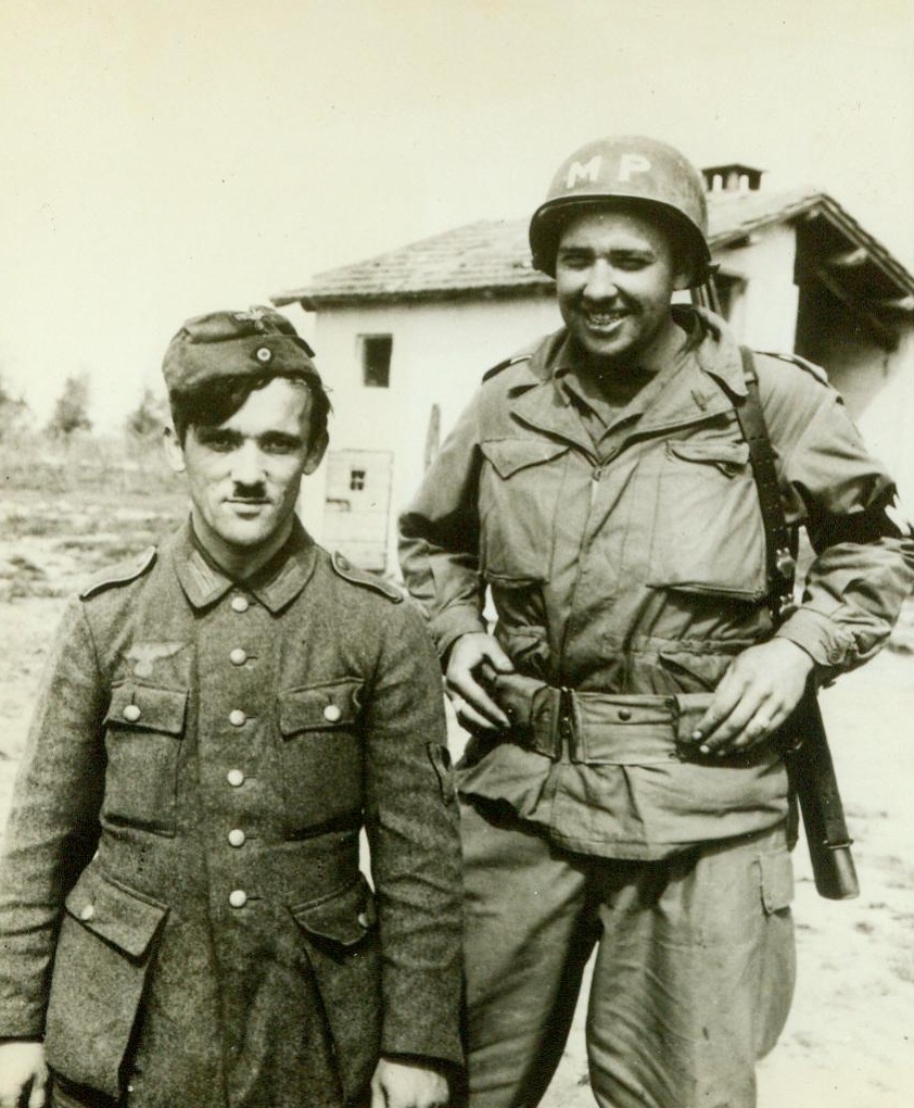 O-OO-OH, That Face!, 5/19/1944. Italy -- This Nazi prisoner, bearing a striking resemblance to Chaplin imitating Hitler poses for the cameraman with grinning M.P. William Spalding, of Dresden, N.Y. Had he not known that Der Fuehrer was safely hiding back in Der Fodderland, Spalding might have thought he had captured the Dictator himself. However, now that the Allies are on the warpath, it won't be long before this phoney picture will be duplicated "in the real" when our forces march into Berlin and out again mit Adolph 5/19/44 (ACME);