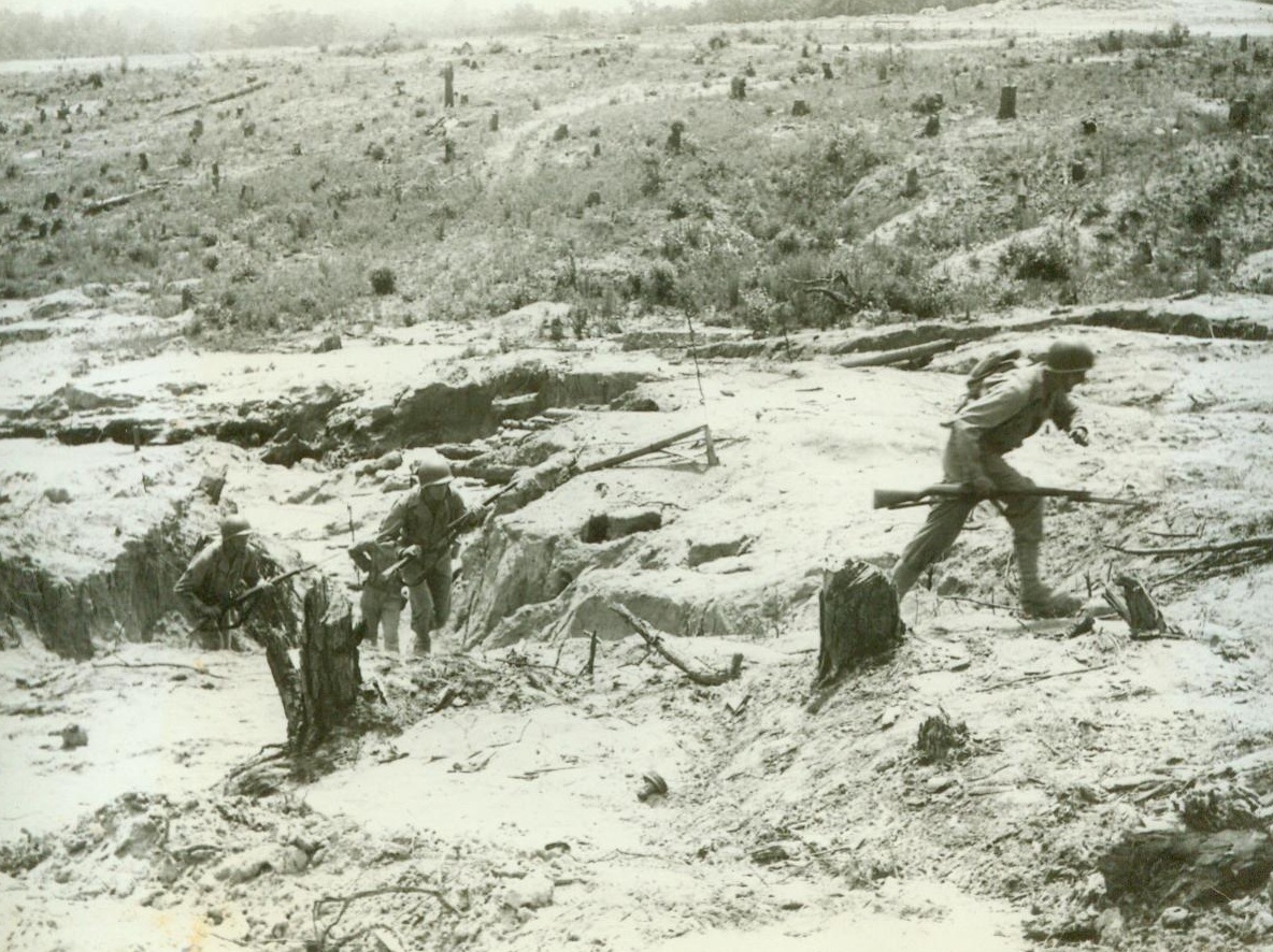 The Infantry Schools West Wall, 5/4/1944. Ft. Benning, Ga. – Newest assault course at Fort Benning is the infantry school’s authentic exterior replica of a section of Germany’s famed West Wall, a series of pillboxes and blockhouses, which extends along the coastline from France to Norway and which our invasion troops must reduce from beachheads before they can drive inland to strike at the heart of Nazidom. Constructed after study from Allied photographs, the course covers 1,000 square yards and is used by the infantry school in the training of officers and officer candidates. Infantry school demonstration troops, some of whom are shown in these photos, carry out “assaults” for the benefit of each new class. Live ammunition is used and every three weeks the concrete pillboxes and blockhouses must be largely rebuilt and barbed wire torn by bangalore torpedoes must be restrung by engineer troops assigned to the school. Photo No. 3: Infantrymen with fixed bayonets advance from ravine and shell holes to support soldier with flame-thrower and to aid in reduction of the pillbox. No make-believe, the box in this picture has a four-foot concrete wall. Note the rugged terrain, the trees reduced to stumps by artillery fire, the pitted earth. 6/4/44 (ACME);