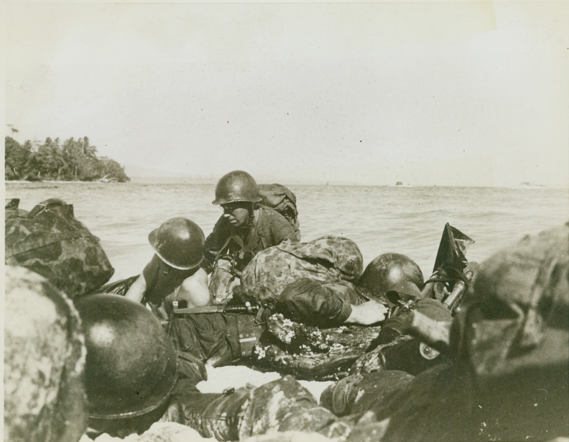 FIRST AID FOR AN INVADER – 3, 6/1/1944. WAKE ISLAND – Fire from Jap machine gun nests and pillboxes streams hotly from the shore, but first aid treatment for the wounded man, who was dragged to this point by his comrades, continues. Fighting man in center has ripped the shirt from his buddy’s back, and begins to dress the wound. Credit: Photo by Lt. Sidney Simon from ACME;