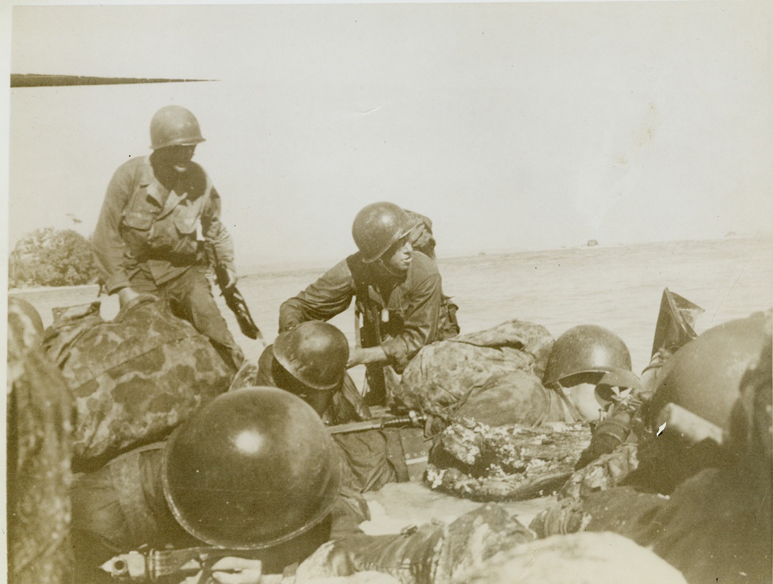 FIRST AID FOR AN INVADER –2, 6/1/1944. WAKE ISLAND – Tense and determined, our infantrymen hug the wet sand as they move through the surf, rifles in hand and full packs strapped to their backs. In center one of the invaders hurries to the assistance of his fallen buddy, stealing a hurried glance toward shore at the same time. Credit: Photo by Lt. Sidney Simon from ACME;