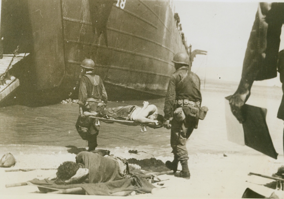 FIRST AID FOR AN INVADER – 5, 6/1/1944. WAKDE ISLAND – The menacing Japanese fire has been silenced now, and medical corps litter bearers have come ashore to collect and evacuate the wounded. The fighting man whose first aid treatment was rendered swiftly and efficiently at the water’s edge lies weakly on the stretcher as it is carried toward the big landing vessel at left. Another felled warrior lies in foreground. Credit: Photo by Lt. Sidney Simon from ACME;