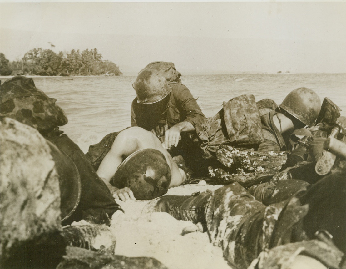 FIRST AID FOR AN INVADER – 4, 6/1/1944. WAKE ISLAND – The wounded fighter has dropped his head to the wet sand, lying there weakly as the bullet holes in his back are cleansed and dressed. Except for the mercy worker, all fighters keep their heads low to avoid the murderous fire from enemy pillboxes and machine gun nests that streams out over the water. Credit: Photo by Lt. Sidney Simon from ACME;