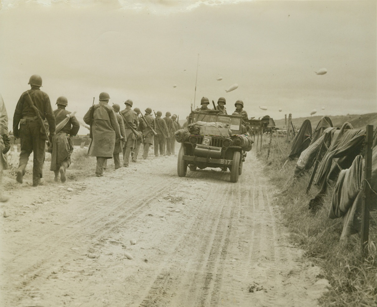 Yanks Headed For Action, 6/13/1944. FRANCE—in single file, American troops march down a narrow French Road on their way from (illegible)…Invasion Beachhead to engage the (illegible)……Note Barrage Balloons, (right,) (illegible)…..(Note:  Some of the left side of the description of this image has been torn away)Credit:  Bert Brandt;