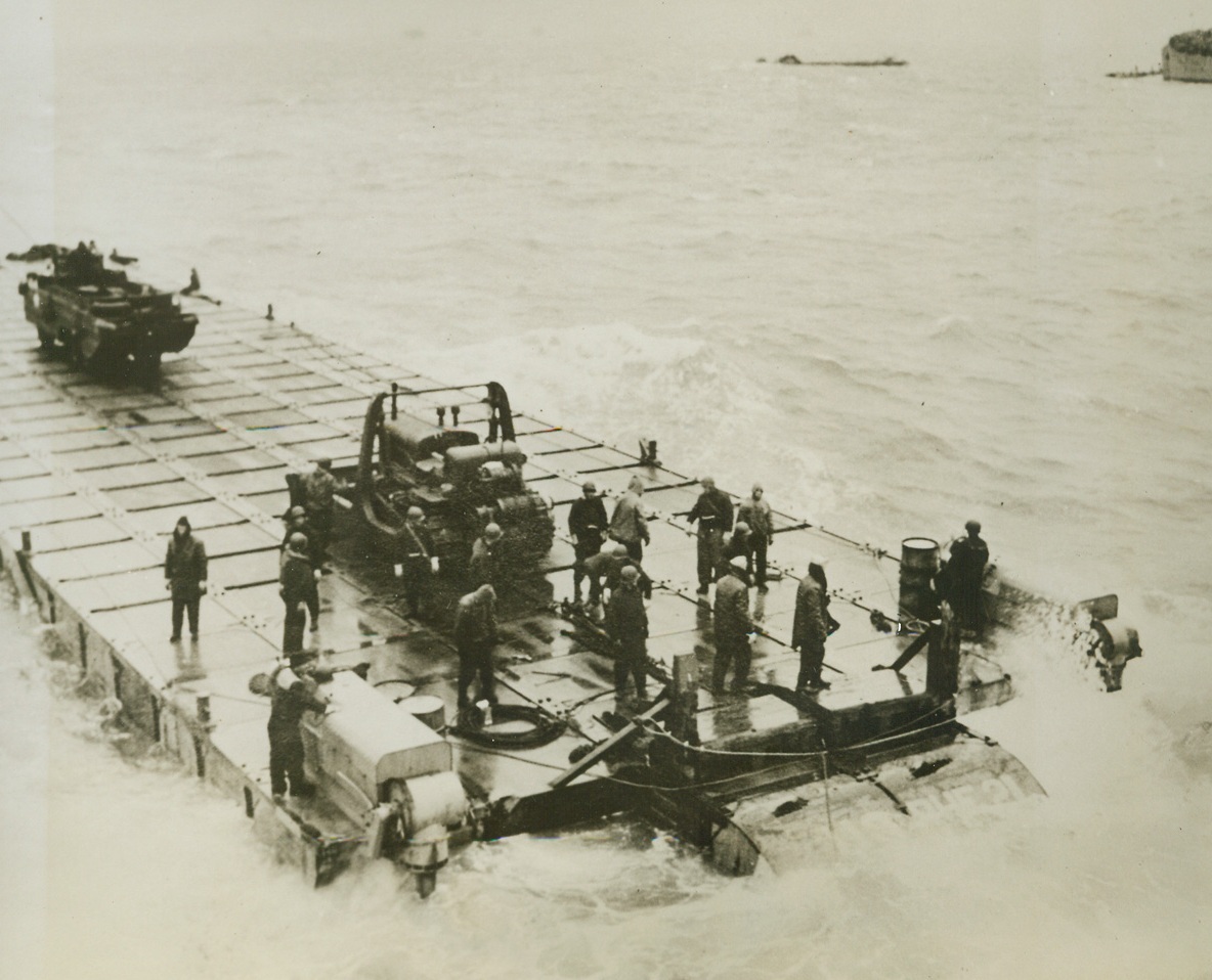 “RHINO FERRY” FOR INVASION FORCES, 6/6/1944. The most difficult phase of the initial operation against the European Continent—that of bridging the last few hundred yards between vessels and the beach—is being accomplished to a large degree through the use of the U.S. Navy’s modern pontoon gear. Made up of hollow boxes of welded metal, in two sizes, a huge, self-propelled barge, called the “rhino ferry” can be put together aboard ship. When the objective is reached, sections of the raft can be lowered over the side, fastened into the whole with bolt, nuts, links, and angles. The, the “ferry” is ready to float vehicles and supplies ashore. The new style pontoons—or tanks—are built in two sizes, five by seven by five feet deep, and the other, seven by seven by five with one edge curved to serve as the prow of a barge. These rafts as a whole—or in sections—can be filled with water and used as piers or docks. Designed by Capt. John N. Laycock, Civil Engineer Corps, USN, Washing, D.C., these pontoons have been used previously in invasions of Attu, Africa, Sicily, and Italy. I this series of photos, the “rhino ferry” is shown being assembled and put to use.  NEW YORK BUREAU Here, the “rhino ferry” gets under way after being assembled. Two powerful Chrysler marine outboard motors at either side of the stern, developing 143 horsepower each, can push the barge along at a little more than four knots. Having a draft less than LSTs, the “ferry” are ideal for the long, shallow beaches of Europe. Credit: U.S. Navy photo from Acm;
