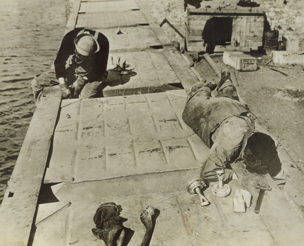 “RHINO FERRY” FOR INVASION FORCES, 6/6/1944. The most difficult phase of the initial operation against the European Continent—that of bridging the last few hundred yards between vessels and the beach—is being accomplished to a large degree through the use of the U.S. Navy’s modern pontoon gear. Made up of hollow boxes of welded metal, in two sizes, a huge, self-propelled barge, called the “rhino ferry” can be put together aboard ship. When the objective is reached, sections of the raft can be lowered over the side, fastened into the whole with bolt, nuts, links, and angles. The, the “ferry” is ready to float vehicles and supplies ashore. The new style pontoons—or tanks—are built in two sizes, five by seven by five feet deep, and the other, seven by seven by five with one edge curved to serve as the prow of a barge. These rafts as a whole—or in sections—can be filled with water and used as piers or docks. Designed by Capt. John N. Laycock, Civil Engineer Corps, USN, Washing, D.C., these pontoons have been used previously in invasions of Attu, Africa, Sicily, and Italy. I this series of photos, the “rhino ferry” is shown being assembled and put to use.  NEW YORK BUREAU Here, two sailors join pontoons together to form the “ferry”. Seabees are given long hours of drill on this assembly until the process becomes almost second nature to them. Credit: U.S. Navy photo from Acm;