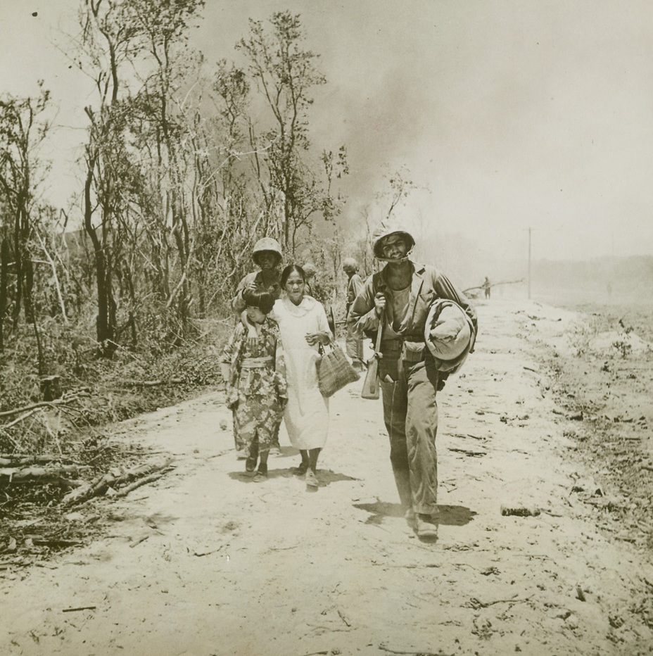 JAP CIVILIANS CAPTURED ON SAIPAN, 6/30/1944. SAIPAN, MARIANAIS—While the Yank in front carries their meager belongings, this Japanese mother and daughter march down the road to the beach where they had to wait until a prison camp was built later in the day. They were captured on the second day of the American invasion of Saipan. In contrast to her mother’s Occidental dress, the little girl wears the traditional Japanese kimono. Credit: ACME.;