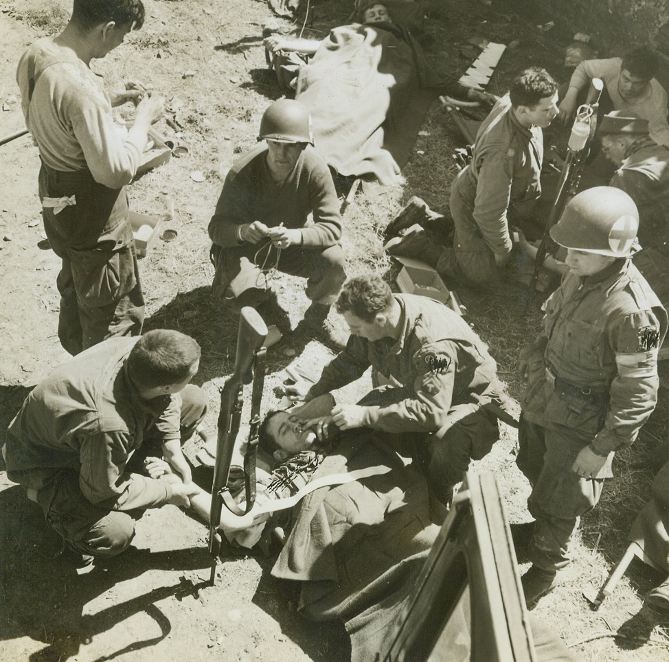 Wounded Warriors, 6/23/1944. St. Sauveur, France—With his rifle stuck in the ground at his side, this American soldier receives medical attention after the battle for St. Sauveur was over, and the town won. In the upper right another wounded man is receiving plasma with the transfusion apparatus stuck on his rifle. Credit: ACME photo by Andrew Lopez, War Pool Correspondent.;
