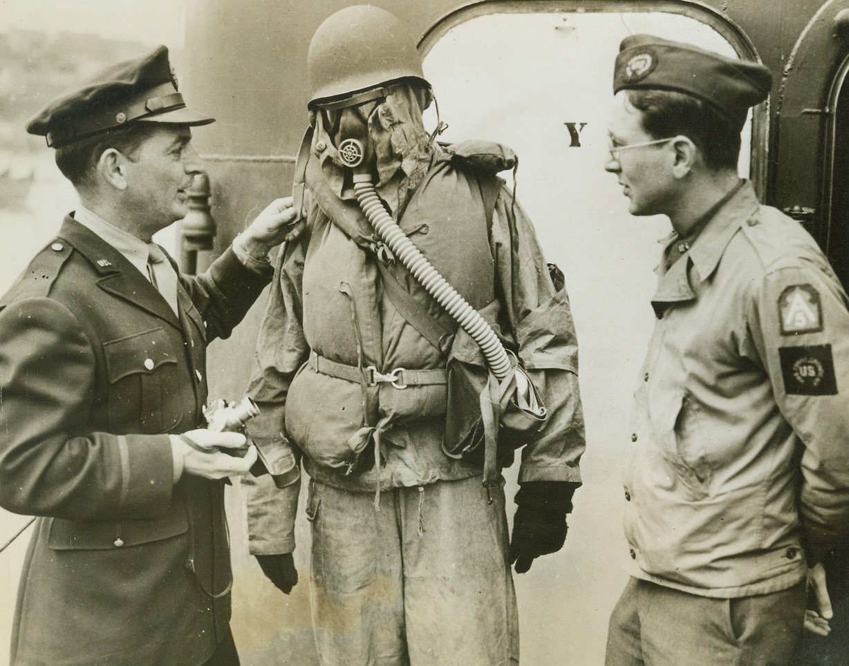 NEWSMAN PREPARES FOR THE WORST, 6/23/1944. AT SEA – Making a personal test to judge how much he will be able to see during combat action in a storm, W.R. Higginbotham of the United Press dons Navy “foul weather gear” aboard a warship.  Jack Rice (left), Associated Press photographer, and Tom Wolf of NEA await his report with interest. Credit: US Navy photo from Acme;