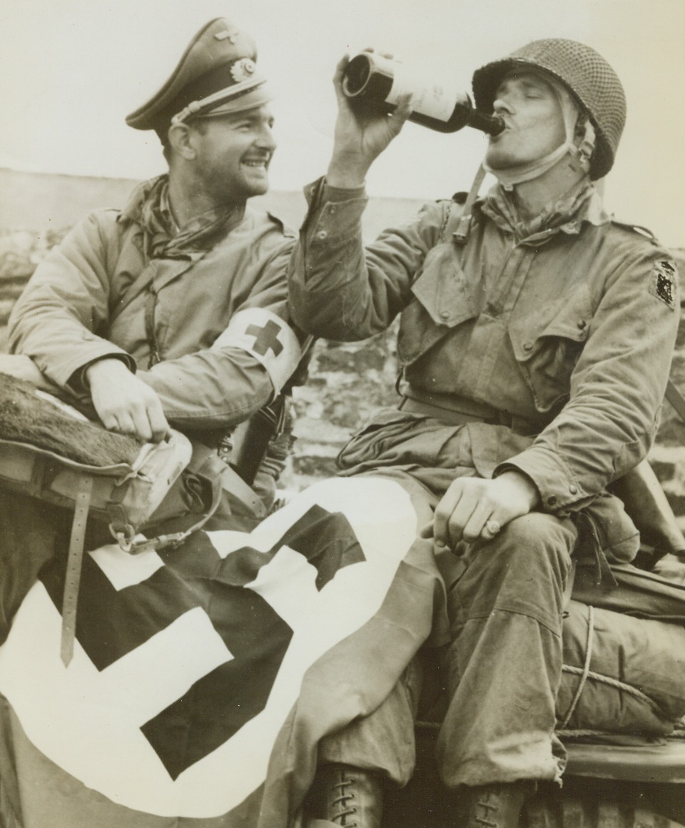 FREED BY ADVANCING YANKS, 6/25/1944. FRANCE – When Lt. Briand M. Beaudin (left), West Warwick, R.I., and Lt. Paul E.K. Lehman, Washington, D.C., parachuted to the ground in France on D-Day, they were captured by German forces in the District of Orglandes.  Yanks advanced into this area and captured it, freeing the two airborne infantrymen.  Wearing a German hat and displaying a Nazi flag, souvenirs of his experience, Lt. Beaudin waits his turn at the wine bottle which the two are sharing in celebration of their liberation.Credit: Acme;