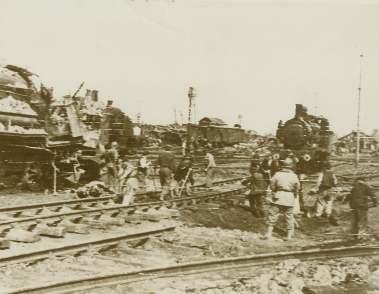 UNDOING THEIR OWN WORK, 6/24/1944. FRANCE – Now that their bombing of French railroads to cut off German supplies and reinforcements has successfully completed its work, American soldiers, with the assistance of French civilians, repair the damaged tracks in the American-occupied area around Cherbourg.  Having surrounded the entire city, Yank troops have placed the key port in a state of siege, and the trapped Germans are fighting desperately.Credit: Signal Corps radio telephoto from Acme;
