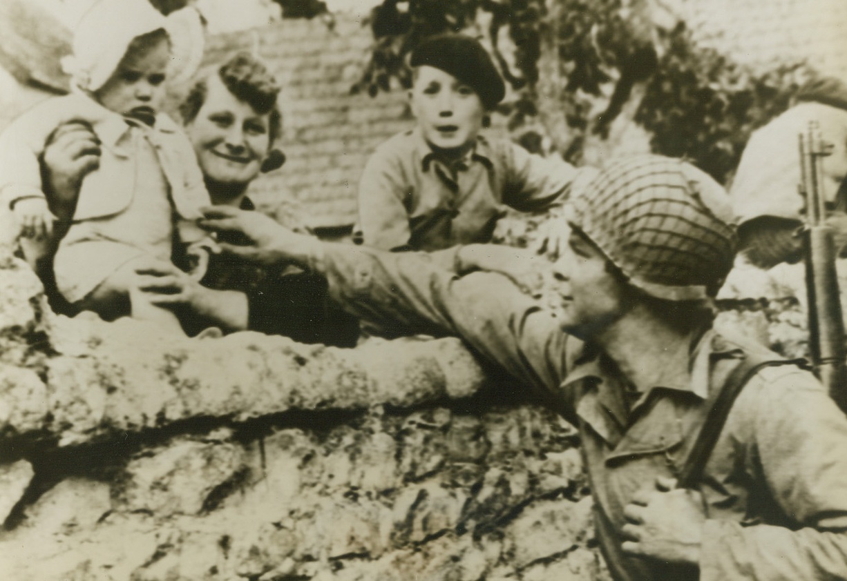 H’Ya Babe, What’s Cookin’?, 6/9/1944. France – With impulsive Yank friendliness, Sgt. Max Denton, of Biloxi, Miss., leans over this stone wall and playfully reaches for the hand of this French youngster while mamma stands by, grinning broadly. Sgt. Denton was with the American Troops advancing into Normandy. Credit: Signal Corps Radiotelephoto from ACME;