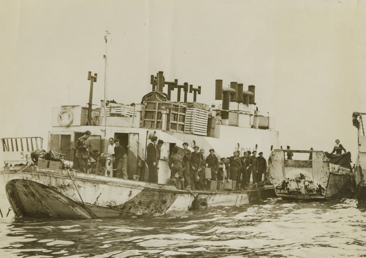 Floating Kitchen, 6/15/1944. France – A converted barge that once sailed up and down the Thames, this floating mess hall was the first Landing Barge Kitchen to reach the Normandy beachhead. Purpose of the vessel is to supply hot meals to the crews of small craft now doing their hundred-and-one jobs in the Channel, which range from ferrying men and supplies to laying smoke screens for air attacks. Here crews of the small craft line up for their noon meal. Credit: British official photo from Acme;