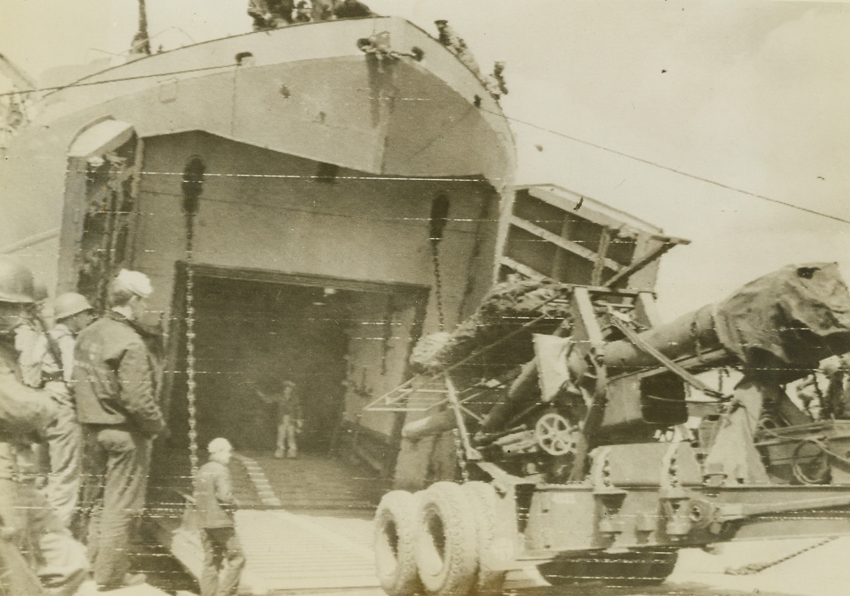 Heavy Stuff for Beachhead, 6/16/1944. England – A huge U.S. gun is backed up the landing ramp of an LST at “a British port”, headed for use on the Allies’ “liberation beachhead” in Normandy. At the top of the ramp a man directs the driver by hand signals. A hint that the Allies expect to capture a deep sea port—possibly Cherbourg—shortly, is indicated by this photo, flashed to the U.S. by radio today. Modern docking facilities would be needed to unload this “heavy stuff.” Credit: Army Radiotelephoto from ACME;