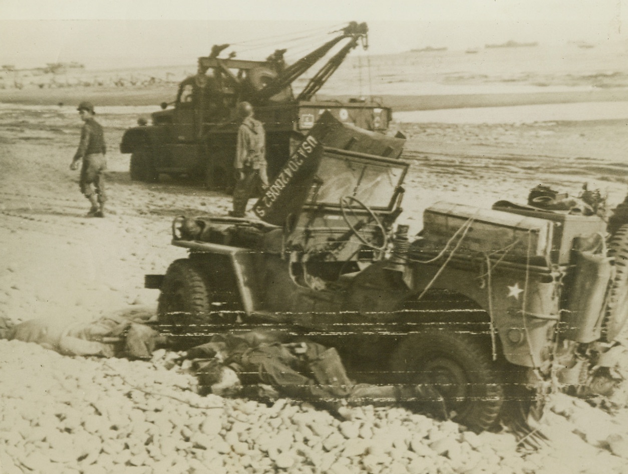 Death—Their Assignment, 6/6/1944. France – Two American soldiers lie dead beside their jeep. They were caught in a deadly German machine gun fire as they rolled down the ramp of their landing craft, and died as they left the tiny car to seek cover. In the background, a prime mover goes about its job of salvaging blasted equipment. Credit: U.S. Coast Guard photo via Army Radiotelephoto from ACME;