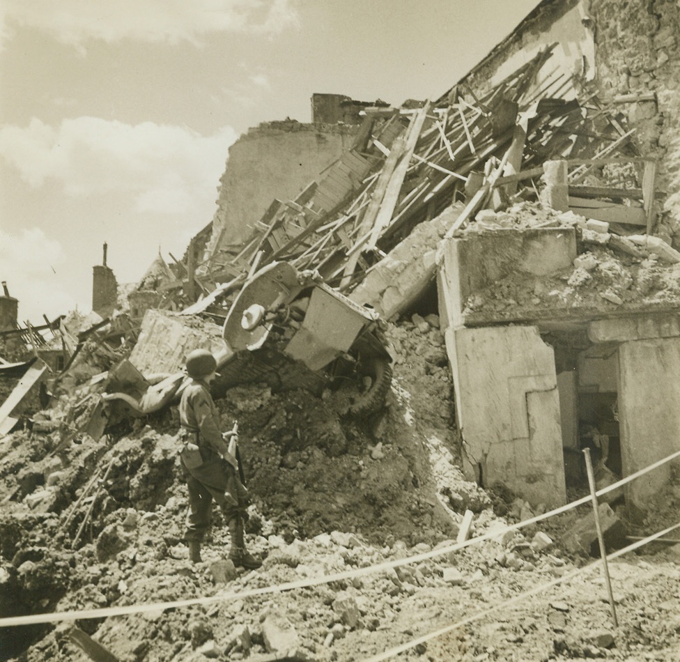 War Passed Here, 6/23/1944. St. Sauveur, France – Standing in an area marked off as “dangerous,” this American soldier looks at the shattered remains of a tank sitting on top of the wreckage of what was once a building in St. Sauveur. American forces have continued their march up the Normandy Peninsula, and are now surrounding Cherbourg, key port on the tip of the peninsula, and their ultimate goal. The capture of the port will give the Allies a good harbor in which to land equipment and troop reinforcements. Credit: ACME photo by Andrew Lopez, War Pool Correspondent;
