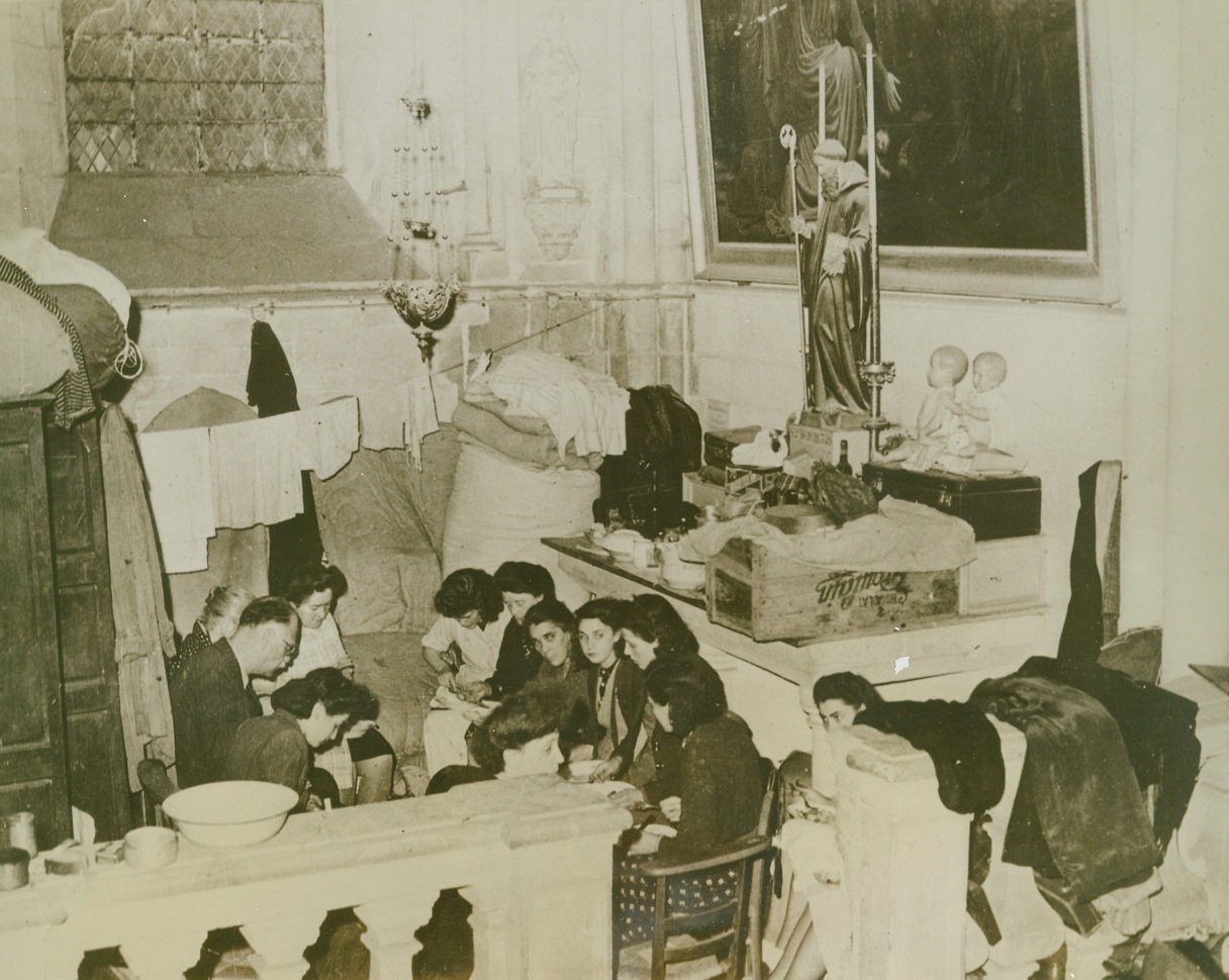 Holy Shelter For Caen Refugees, 7/18/1944. CAEN, FRANCE—Civilians  take refuge in the cathedral at Caen.  One of the alters is used as a supply table with a clothesline running in front of it and a refugee family has it’s meal in one of the alcoves of the church.  Note the two dolls reposing on the alter.Credit (ACME);