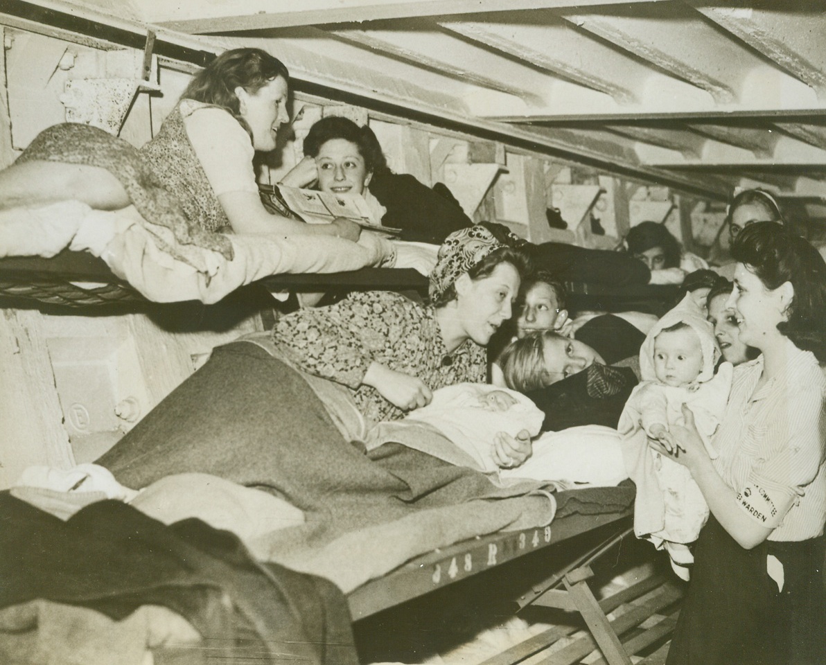 New Deep Shelter Against "Buzz Bombs", 7/27/1944. London, Eng. – TO allow Londoners protection and to give them some opportunity to sleep while the city is under bombardment by German robot “buzz-bombs”, the first of a series of deep shelters has been opened. These new shelters consist of two main twin tunnels running beneath existing tube railway (subway) lines. They are divided into 16 sections each fitted with bunks for 500 persons. Cross-passages connecting the two tubes contain first aid posts, lavatories and machinery control rooms. Bunks fold against the walls when not in use to allow free passage of traffic. Four canteens are provided at suitable points, run by volunteer organizations. The shelters are controlled and administered by commissioners for the London Civil Defense Region. Each shelter is in charge of a shelter superintendent, with a nucleus staff of full-time wardens, supplemented by volunteer wardens. In one of the sections of the new deep shelter, Londoners lie in the double row of bunks, at the right, a warden holds a child. Credit ACME;