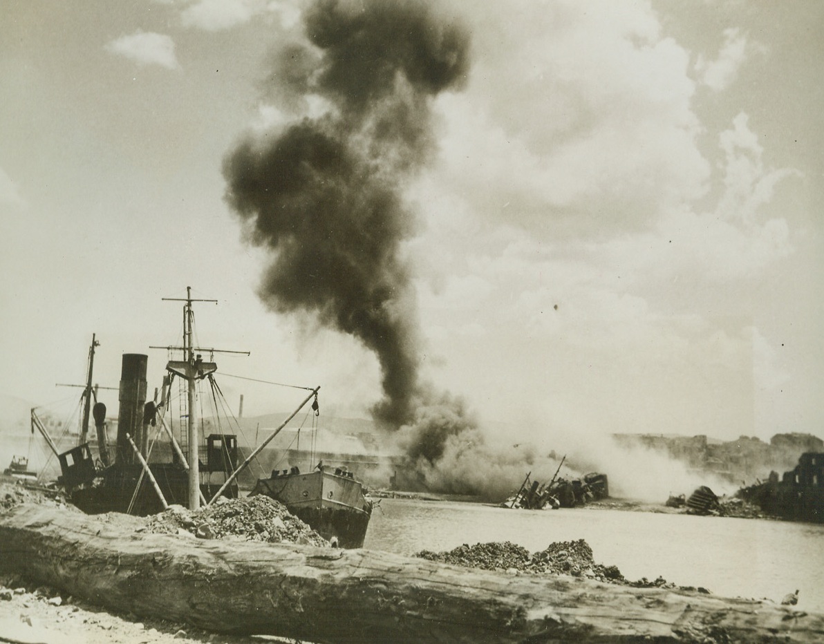 Make Way for Allied Supplies!, 7/3/1944. CIVITAVECCHIA, ITALY—Smoke rises above the harbor at Civitavecchia after American troops blew up a beached ship with a hearty dose of TNT. The ship was blocking the harbor entrance and preventing LST’s to land with supplies. Allied troops are steadily advancing toward the Pisa-Rimini Line, with French Forces with the 5th Army capturing the historic city of Siena, and Americans advancing past Cecina toward Leghorn.  Credit:  ACME;