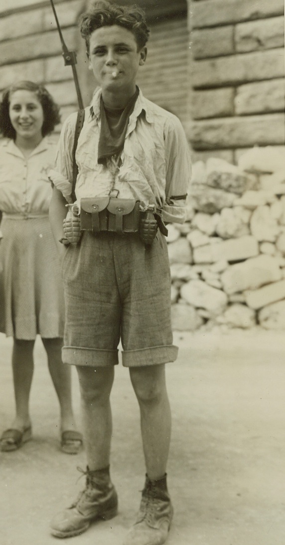 PINT-SIZE PARTISAN, 7/24/1944. LIVORNO, ITALY—Puffing nonchalantly on a cigarette and carrying a man-sized rifle, two hand grenades, and other arms, this youthful partisan presents an incongrous [sic] picture. Familiar with the layout of Livorno, he directed the American troops away from heavily-mined areas.Credit: Acme photo by Sherman Montrose, War Pool correspondent;