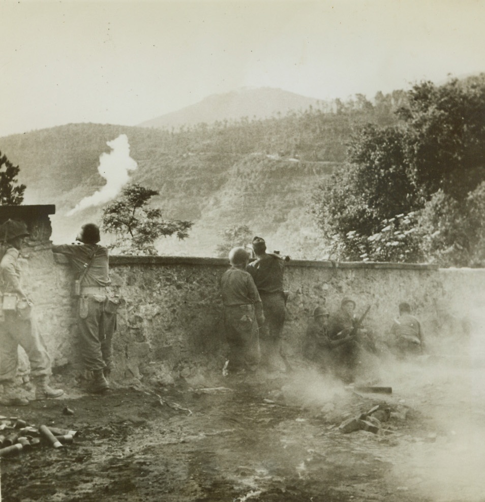 Yanks in a Spot – 4, 7/14/1944. Nemi Valley, Italy – Once the peaceful site of fragrant vineyards and the center of a flourishing wine-grape business, beautiful Nemi Valley, near Rome, was turned into a hot spot recently when a scouting patrol of Yank Infantrymen, guided by Italian partisans, found German strength much greater than they had suspected. Our boys had to fight their way out through holes in a wall of enemy machine gun fire. Now the tables are turned and the Yanks, protected by a stone wall, are dishing it out to enemy troops in the valley below. Credit: Yank Magazine photo from ACME;