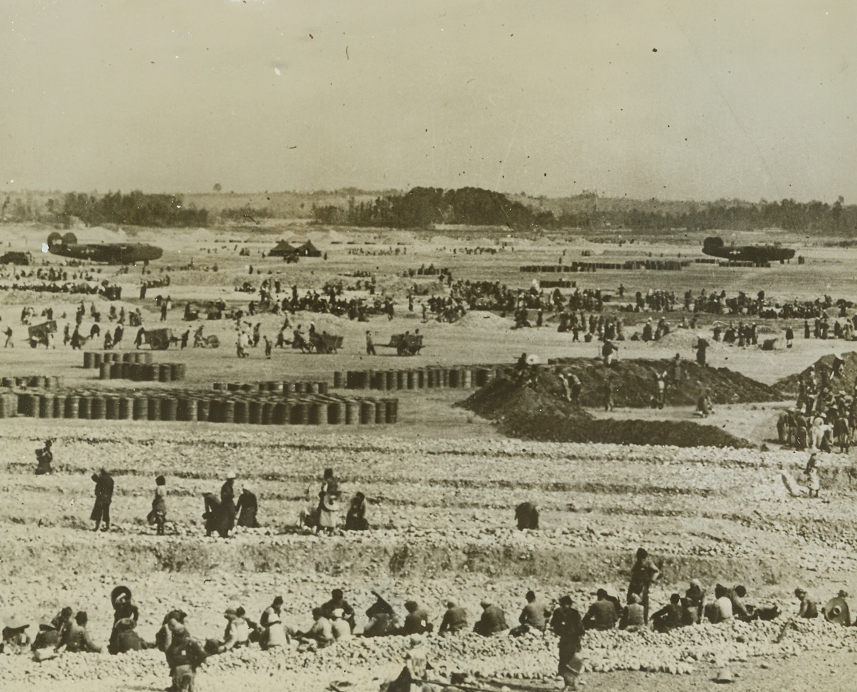 Home for Super Forts, 7/16/1944. This field in Western China is a beehive of activity as Chinese peasants cart up river-bed stone to be used in the construction of an airbase for 20th Bomber Command Superfortresses. The stone is crushed by the only machine used in the construction work. It will be used to cover the landing strips. Note the large number of oil and fuel drums in the center. C-47 transport planes in the background landed before the field was completed.Credit: ACME;