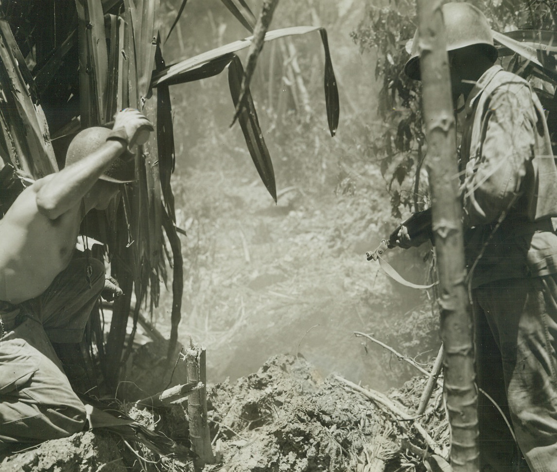 Blasting Out Dug-In Japs, 7/12/1944. Saipan – Meeting increased resistance from Japs on the Northern tip of Saipan, American soldiers are shooting, bombing and smoking the enemy from caves and deep fox holes, such as this one.  The Yank at left is tossing another smoke bomb into the hole to force it’s reluctant occupants out in the open as the rifleman at right prepares to finish them off if they come out fighting.Credit Line – WP – (ACME photo by Stanley Troutman, War Pool Correspondent);