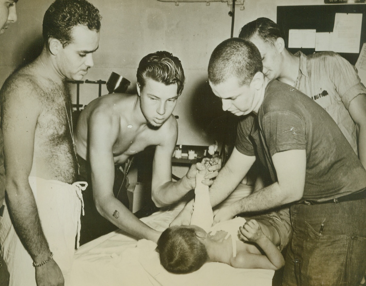 GENTLE HANDS TO HELP HIM, 7/9/1944. SAIPAN—Tenderly, Coast Guardsmen dress the wounds of a young Japanese boy, an innocent bystander who suffered serious wounds in the battle for Saipan. Forgetting the child’s nationality, the fighting men provide medical attention in the American way, aboard a Coast Guard manned transport. Left to right: Jack Schwartz, PhM 3/c, Atlantic City, N.J.; Arnold Allen, S l/c, Portland, Oregon; and Charles Schlicter, PhM 1/c, Harrisburg, Pa. Credit: U.S. COAST GUARD PHOTO FROM ACME.;
