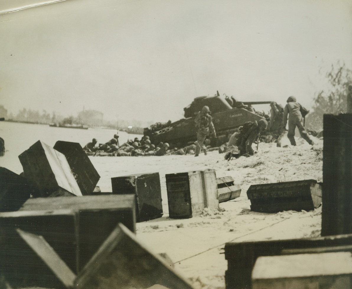 DUCK FOR COVER, 7/10/1944. SAIPAN—Members of a Marine assault wave duck behind a tank for cover as the Japs spray the beachhead at Saipan with machine gun fire. The Leatherneck on his knees in center of photo appears to have been hit while scrambling for shelter. Credit: MARINE CORPS PHOTO FROM ACME.;