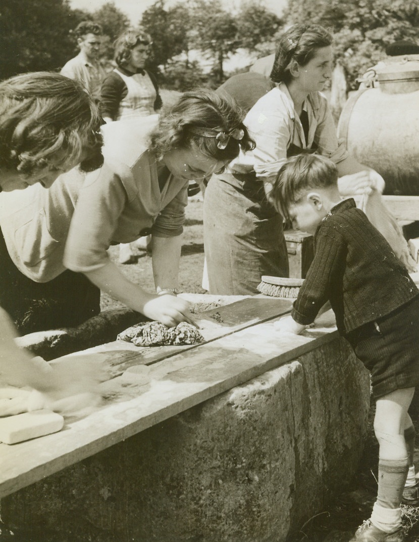 Mother’s Little Helper, 7/10/1944. Normandy, France—Intent upon his work, this little French boy scrubs away as he gives a hand to French women refugees as they do their washing. Note the old-fashioned way they wash—using boards to scrub the clothes on, just as their ancestors did in days gone by. They are in special camps for refugees established by Allied civil affairs. Credit: ACME photo by Andrew Lopez, War Pool Correspondent.;