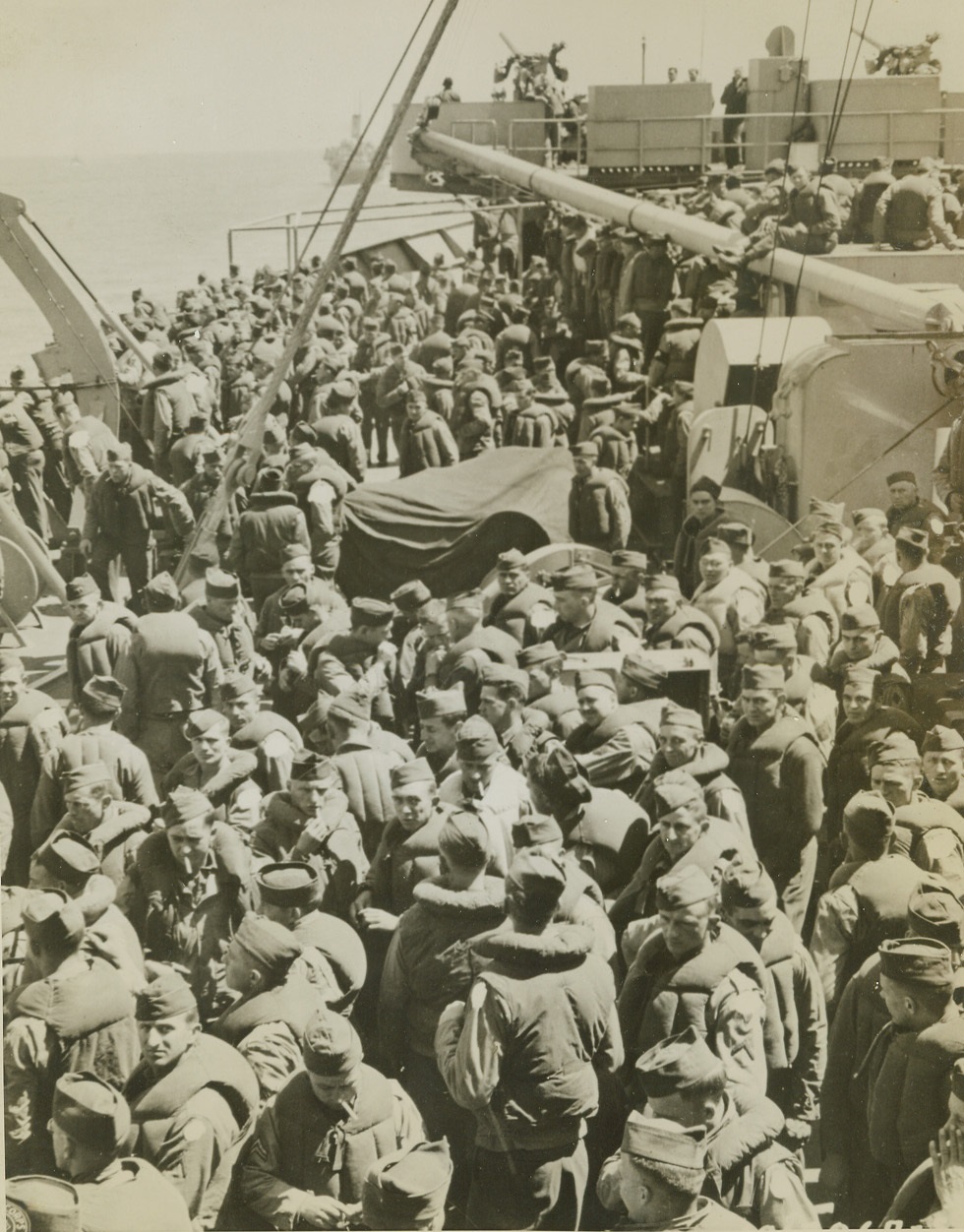 They’re Going Over,  7/17/1944. En Route to France—Bundled up in puffy life jackets and crowding together on the deck of an Allied vessel taking them to the coast of France, American infantrymen smoke and chat about the adventure before them. They’ll reinforce Yank troops now chasing Jerry back across the fields of France.  Credit: Signal Corps Photo from ACME.;