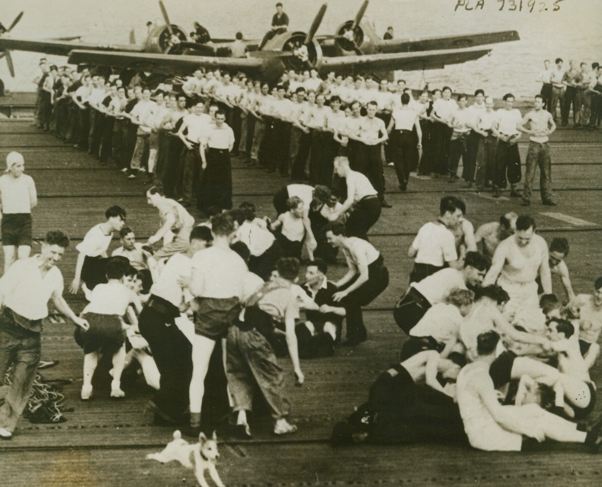 Just a Friendly Tussle, 7/28/1944. The crew of the British Escort Carrier HMS Searcher keep in physical trim by staging a free-for-all tussle on the ship’s flight deck. “Able Dog Spud”, the ship’s mascot, (foreground) flees from the mad scramble, while the rest of the crew (background) awaits its turn. Credit: British official photo from ACME;