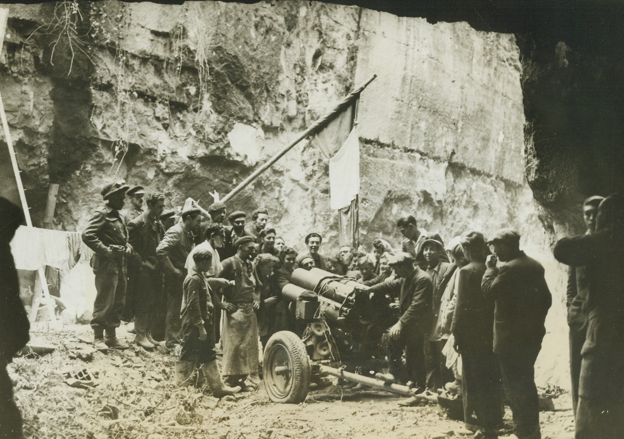 “MOANING MINNIE” IS KAPUT, 7/26/1944. FLEURY-SUR-ORNE: Patriots raise their French flag over a six-barrelled German mortar, which was called “moaning Minnie” and which had been posted at the mouth of a cave near here.  The cave had also been used by the Germans for storing ammunition, and for the last six weeks had been used as a shelter for 900 people of the village. Credit: Acme;