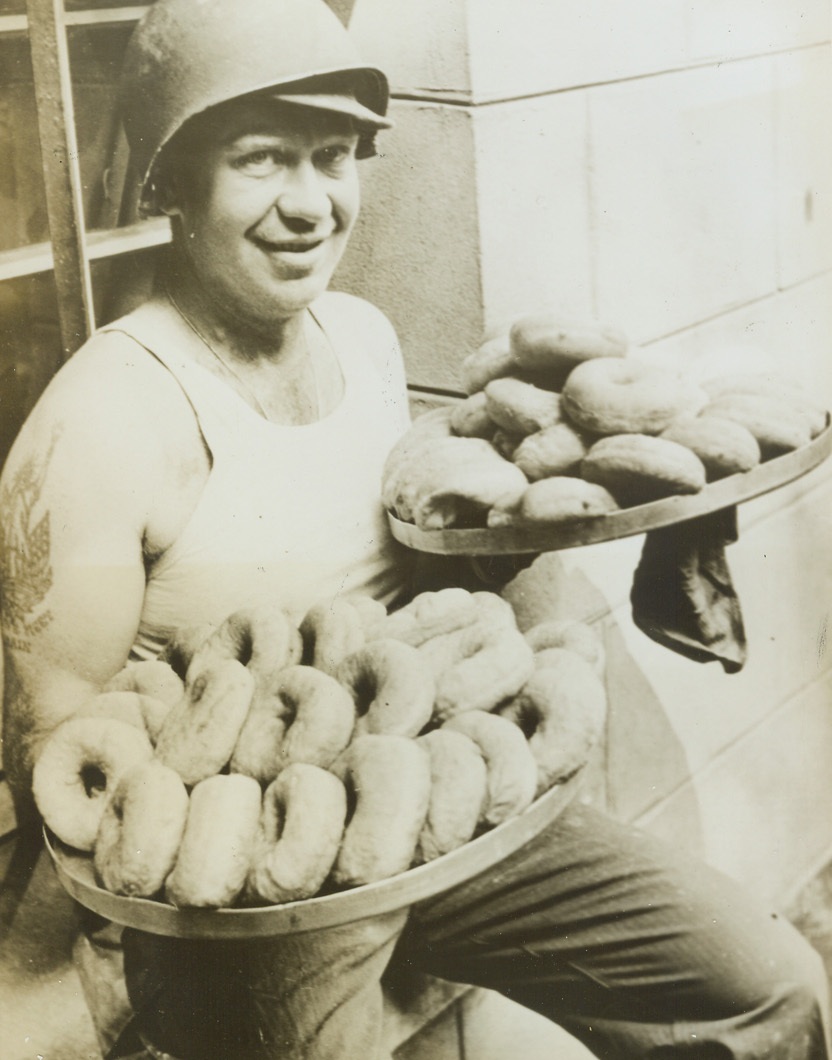 SINKERS FOR THE JAVA, 7/15/1944. LA HAYE DU PUITS, FRANCE—With a sly grin on his face and a twinkle in his eye, Pvt. Paul Taylor, Springfield, Mo., Division Artillery H.Q. mess cook, pauses enroute with a surprise for the ever-hungry Yank forces in his division. His peculiar knack with a few simple ingredients has produced two lovely platters of raised doughnuts, and with a little java, the boys have a great treat in store for them. Credit (ACME) (WP);