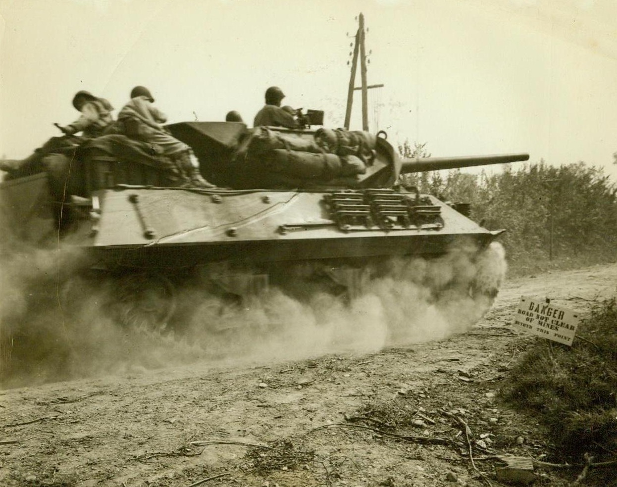 Moving Up For The Kill, 7/24/1944. St. Lo, France - An American Heavy Tank, loaded with Infantrymen, sweeps into the outskirts of St. Lo for the final attack on the town. Note sign on roadside at right: “Danger, Road Not Clear of Mines Beyond This Point.” Credit: ACME photo by Andrew Lopez, War Pool Correspondent;