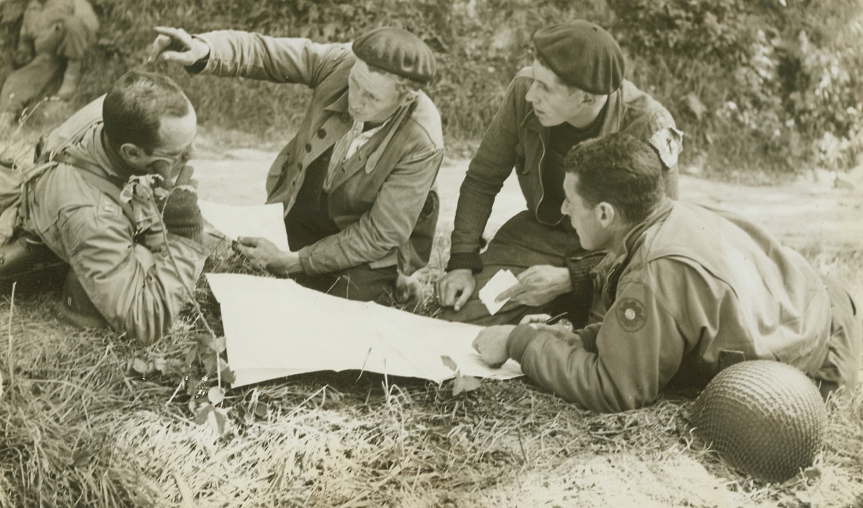 The Underground Has Its Day, 7/17/1944. Normandy – After four long years of fearless resistance to the Nazi oppressors of France, the French Underground today is gradually emerging from hiding to fight the last lap of its battle for freedom. Gleaning vital information about German fortifications, the patriots try to establish contact with Allied forces moving through Normandy. They come in small groups of two and three, bringing their useful news to Allied commanders, sometimes revealing enough Nazi secrets to shorten our battle by days, even weeks. In these first photos of patriots joining hands with Yank forces, two Frenchmen make their report at American Headquarters near Cherbourg, before that city fell to the Yanks. One aged 18, and the other aged 27, the patriots had spent a day behind German lines defending that city before making their way to American Headquarters. A map of the surrounding terrain spread out before them on the ground, the French patriots point out the exact location of German defenses in the Cherbourg area. The men also revealed information on emplacements, troop concentrations and troop movements to Lt. Carl Ruff (extreme right), of New York City, and the Captain at left. Credit: ACME photo by Bert Brandt, for the War Picture Pool;