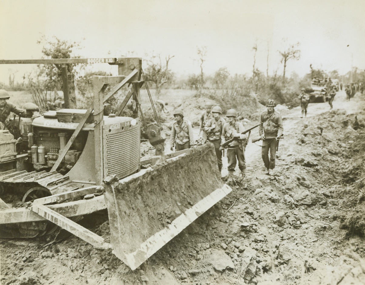 It Puts In and Takes Away, 7/29/1944. France – While trucks wait in the background, the mighty bulldozer fills in bomb craters on a road in France, to clear the way for advancing American forces. The versatile machine has also proved a great asset in clearing debris-filled streets in captured towns. Credit: Signal Corps photo from ACME;