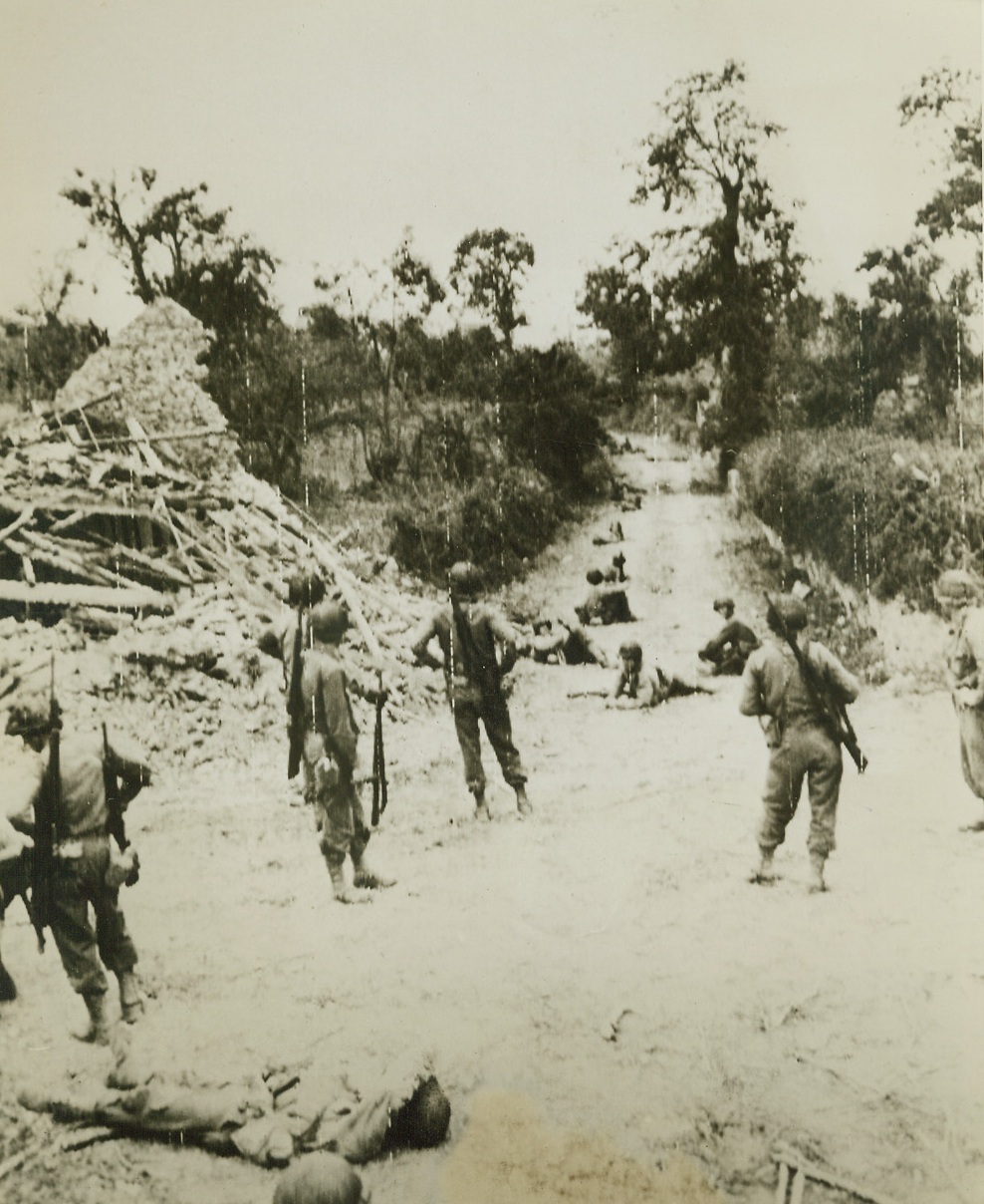 Yanks Stop for Chow, 7/20/1944. France – Beside a wrecked building north of St. Lo, relentlessly advancing American infantry men pause for chow before pushing on to their next objective. Soldiers line the road leading past the ruined house, sitting, reclining, and some catching forty winks. Credit: Signal Corps Radiotelephoto from ACME;