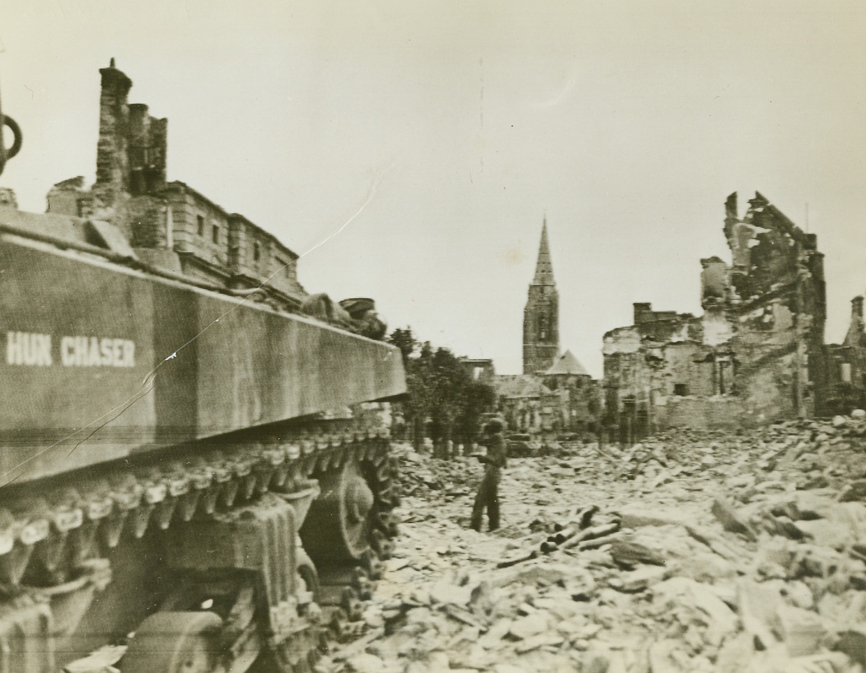 “Hun Chaser” on the March, 7/21/1944. St. Lo, France – While the gigantic “Hun Chaser” tank rumbles through the debris of St. Lo, Pvt. Walter Hatfield, Princeton, Idaho, looks for snipers with field glasses and poised pistol. Blockbusters have not yet cleared the streets of rubble. Credit: US Army Radiotelephoto from ACME;