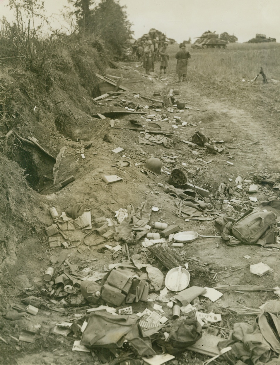 “High Water Mark” of Battle, 7/25/1944. St. Lo, France – Abandoned foxholes and hurriedly discarded effects mark a line behind this hedgerow where American troops rested before plunging on into the battle for St. Lo. Helmets, playing cards, mess kits, and hundreds of other belongings litter this field. In the background, tanks and men are gathered in the field. Passed by censors. Credit: ACME Photo by Bert Brandt for War Picture Pool;