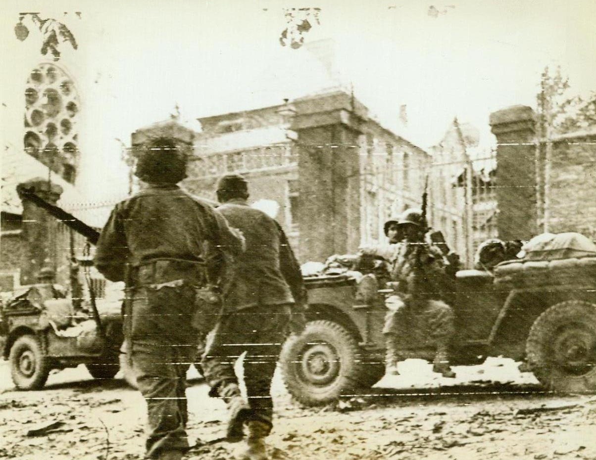 Nazi Captured Under Fire, 7/20/1944. France -- An American soldier (left, foreground) rushes a captured German through the town square in St. Lo, even as the spot was being blasted by enemy artillery. The yank in the jeep at right, points and shouts instructions to the man and his prisoner. 7/20/44 (ACME);
